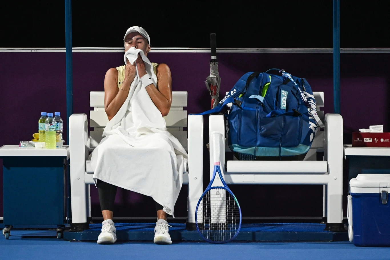 Magda Linette Zawodniczka siedząca na ławce na korcie tenisowym, okryta ręcznikiem, wycierająca twarz, obok niej duża sportowa torba, rakieta tenisowa oraz butelki z wodą.
