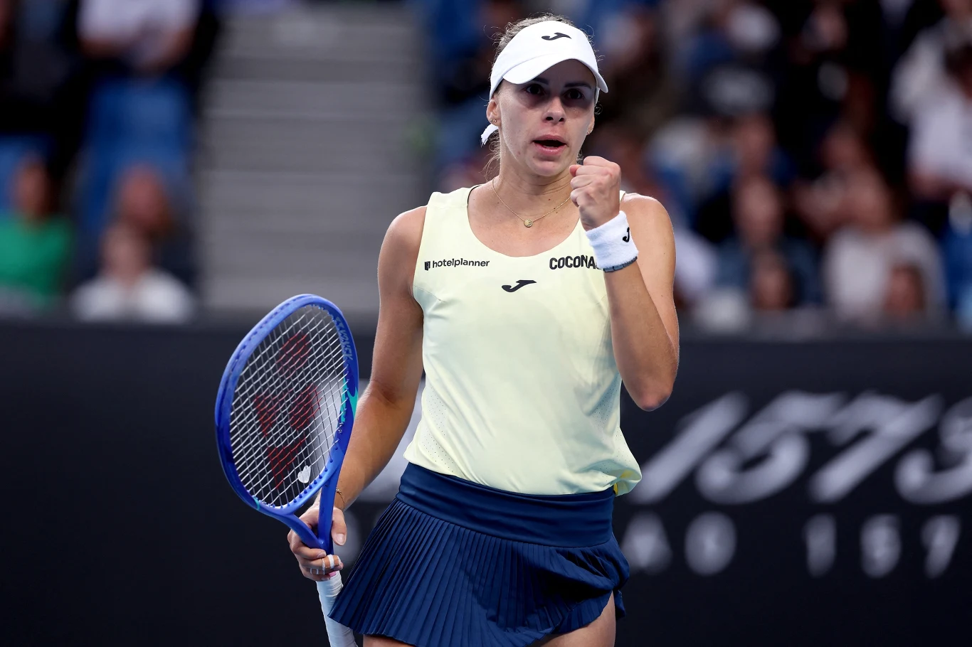 Magda Linette Tenisistka wykonuje gest triumfu podczas meczu na korcie, trzymając rakietę w lewej ręce, ubrana w biały top i granatową spódniczkę sportową, w tle widoczna rozmyta publiczność.