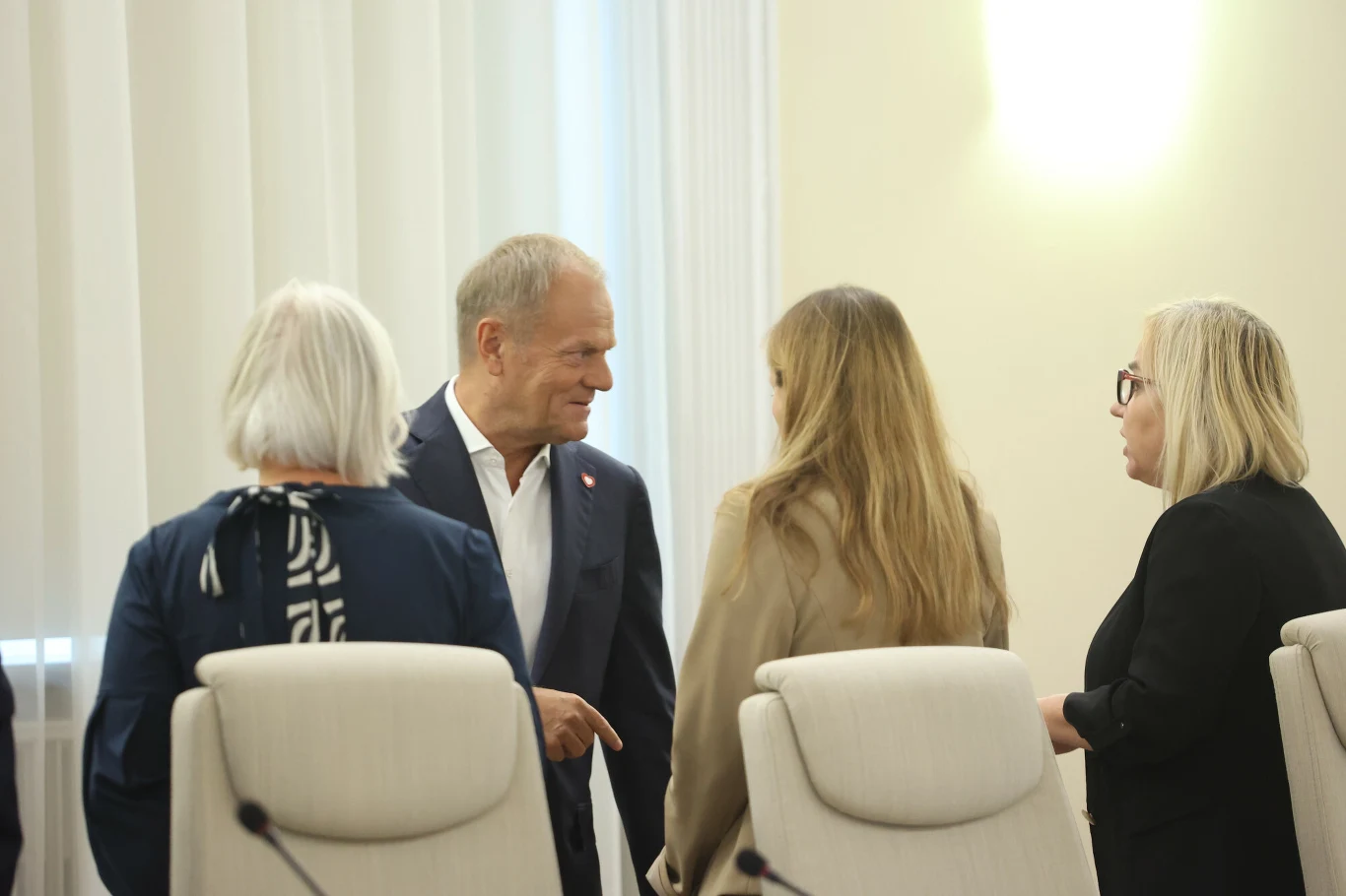 Donald Tusk komentuje rozpad Polski 2050. Na zdj. premier z Katarzyną Pełczyńską-Nałęcz i Pauliną Hennig-Kloską Grupa osób rozmawiających ze sobą w sali konferencyjnej, mężczyzna w garniturze uśmiecha się i prowadzi rozmowę z trzema kobietami, wszyscy stoją wśród jasnych krzeseł, w tle białe zasłony i jasna ściana.