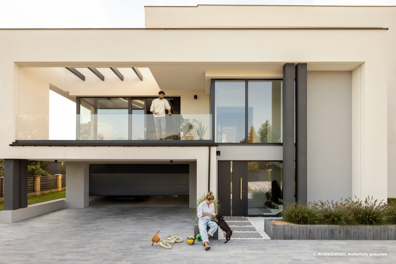 Nowoczesny, piętrowy dom z dużymi przeszkleniami, balkonem oraz przestronnym podjazdem. Na tarasie znajduje się mężczyzna, natomiast na podjeździe kobieta siedzi z dzieckiem i psem, a obok rozłożone są zabawki. Otoczenie zadbane, z fragmentem zieleni p...