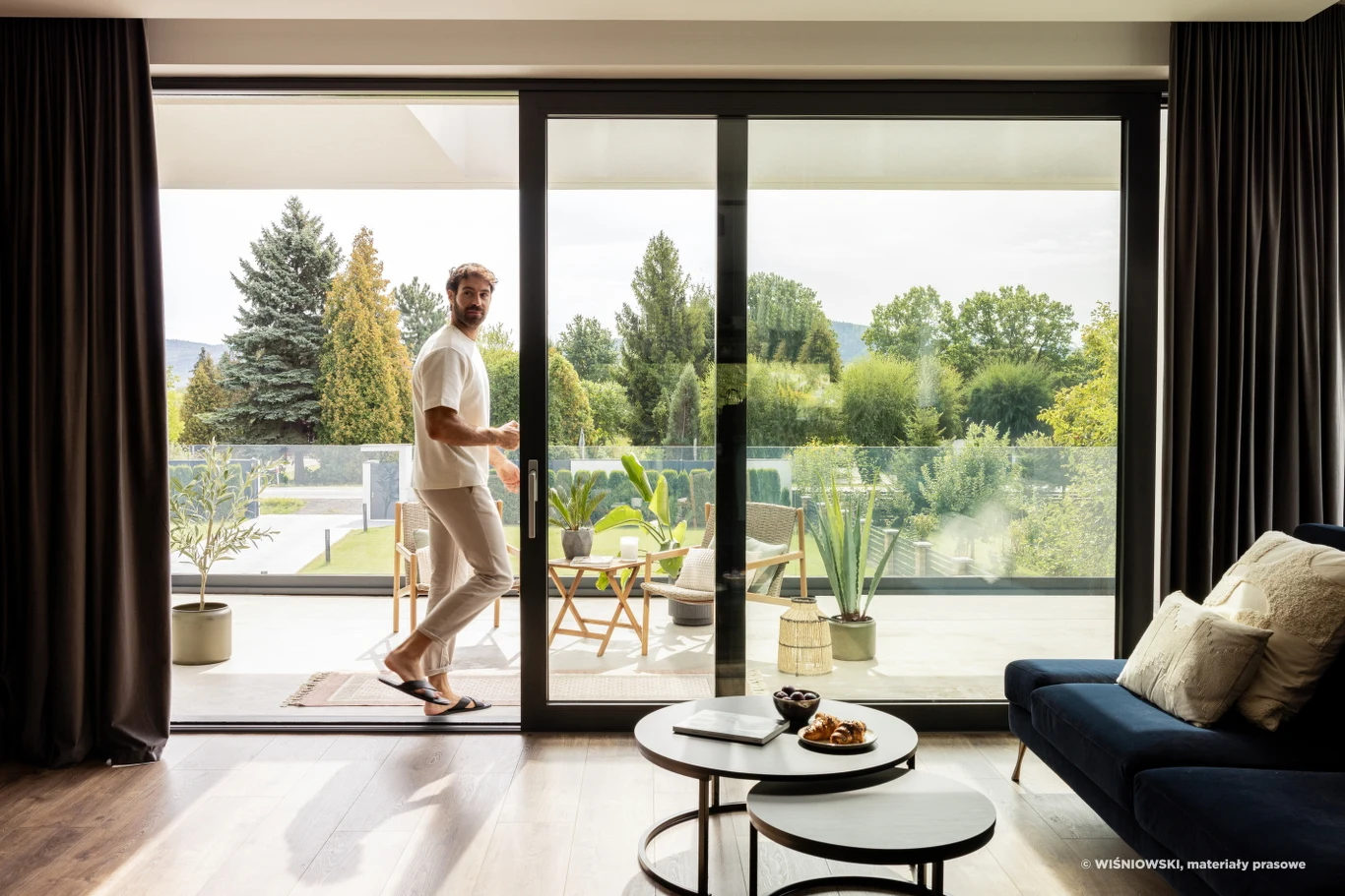 Salon z dużymi, przeszklonymi drzwiami prowadzącymi na taras, przez które przechodzi mężczyzna w jasnych ubraniach. Na tarasie rośliny doniczkowe i widok na zieleń oraz drzewa, w tle wygodne meble ogrodowe. W środku widoczna kanapa i stoliki kawowe z p...