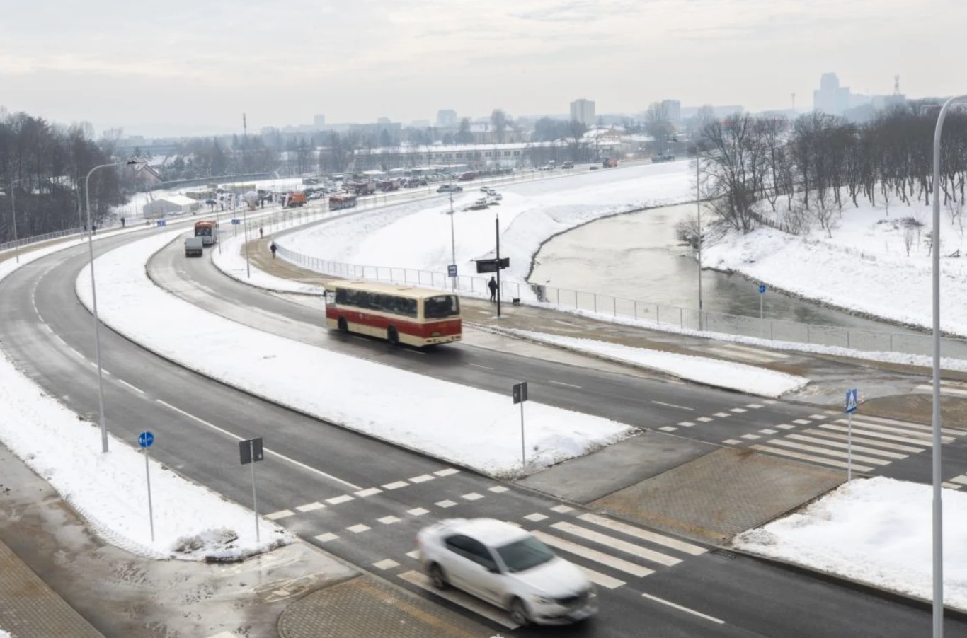 Zakole ruchliwej drogi, po której poruszają się samochody i autobus, otoczone zaśnieżonym terenem w pobliżu rzeki, w tle widoczne są zabudowania miejskie oraz drzewa.