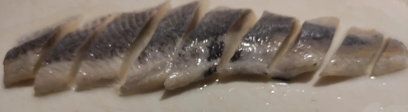 Pokrojony filet śledziowy ułożony na jasnym podłożu, kawałki ryby mają srebrzysto-szarą skórę i miękko połyskującą strukturę.