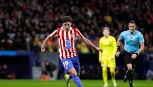 Zawodnik drużyny w czerwono-białych pasach prowadzi piłkę po murawie podczas meczu piłkarskiego, w tle widoczni inni piłkarze oraz sędzia.