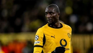 Piłkarz w żółto-czarnym stroju sportowym podczas meczu, na tle rozmytej publiczności i stadionu z wyraźnie widocznym logotypem sponsora na koszulce.