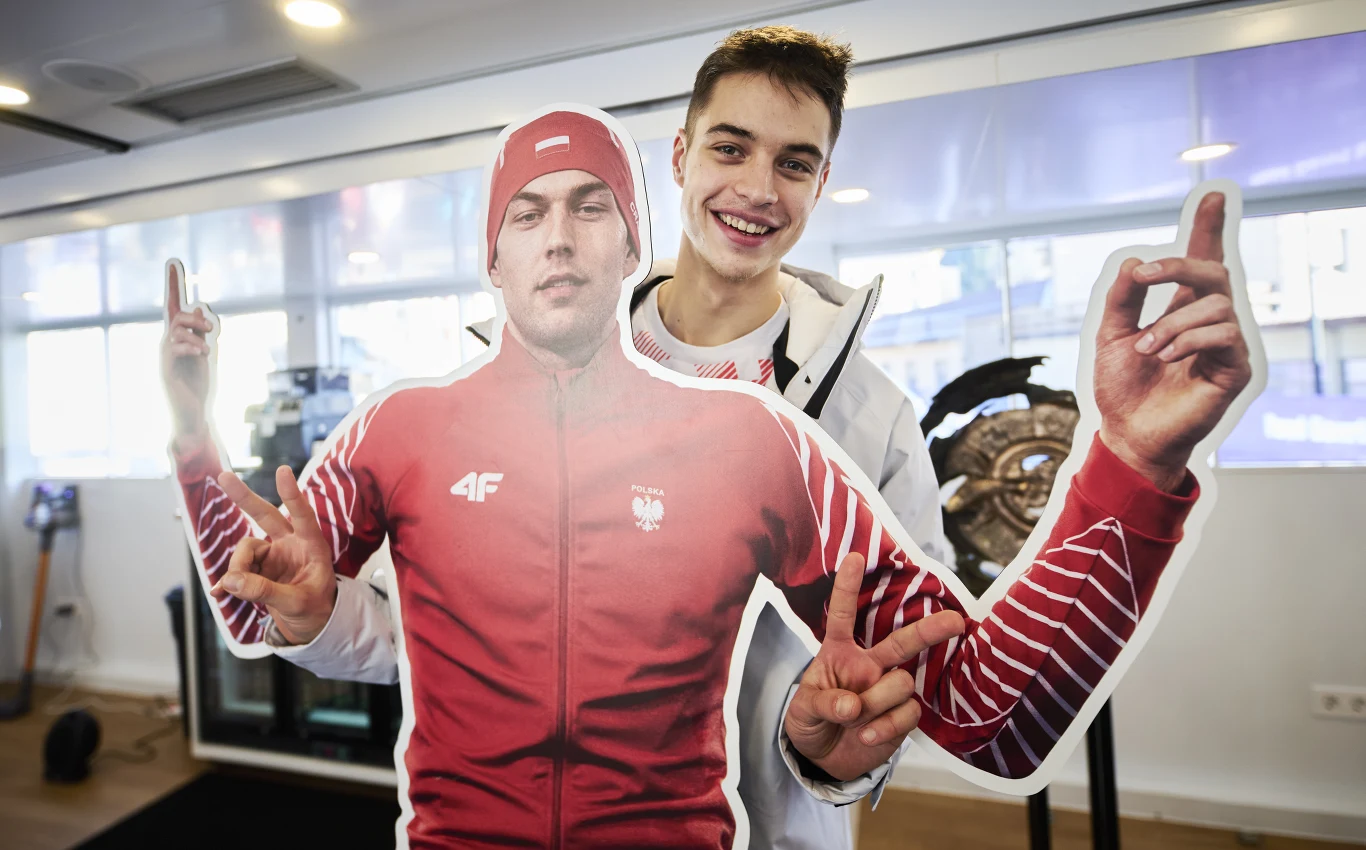 Władimir Semirunnij Młody mężczyzna w białej kurtce i czerwonym swetrze pozuje uśmiechnięty za kartonowym standem wizerunkowym sportowca w czerwonym stroju reprezentacyjnym z orłem na piersi, unoszącym obie dłonie z wyciągniętymi palcami wskazującymi.