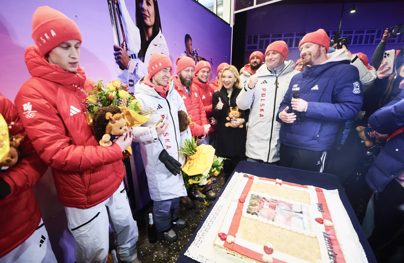 Polscy skoczkowie w domu polskim Grupa sportowców ubranych w biało-czerwone stroje i czerwone czapki celebruje osiągnięcie przy dużym torcie oraz bukietach kwiatów, otoczona przez kibiców i obsługę, panuje radosna atmosfera.