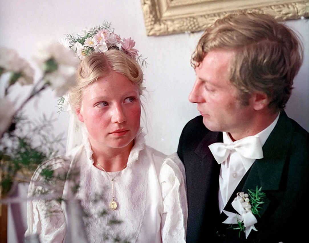 "Pójdziesz ponad sadem" Młoda kobieta w białej sukni ślubnej, z kwiatami we włosach, siedzi obok mężczyzny w czarnym garniturze i białej muszce. Oboje mają poważne wyrazy twarzy, wokół widoczne są rośliny oraz fragment ozdobnej ramy na ścianie.