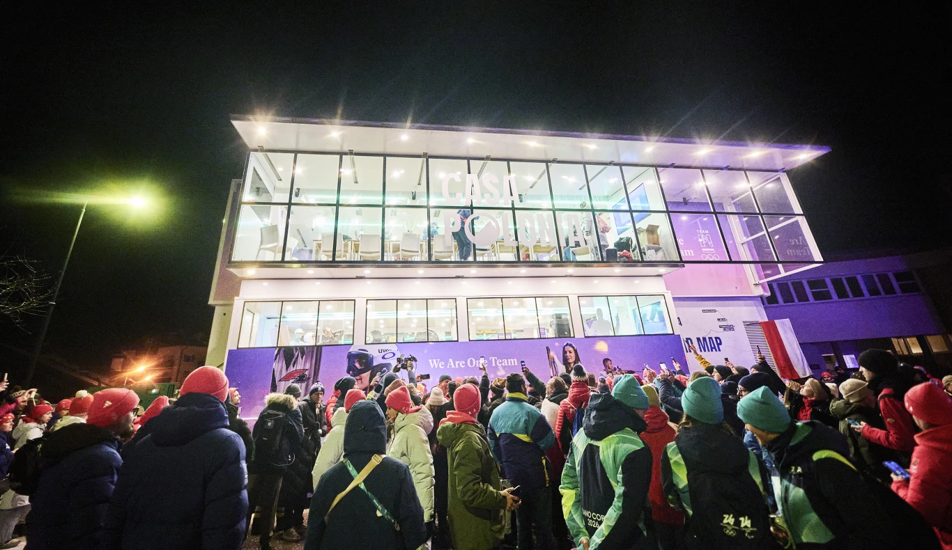 Dom Polski Tłum ludzi zgromadzony przed nowoczesnym, szklanym budynkiem w nocy, wiele osób ubranych w kolorowe kurtki i czapki, budynek jest oświetlony i widać w nim ludzi także na piętrze.