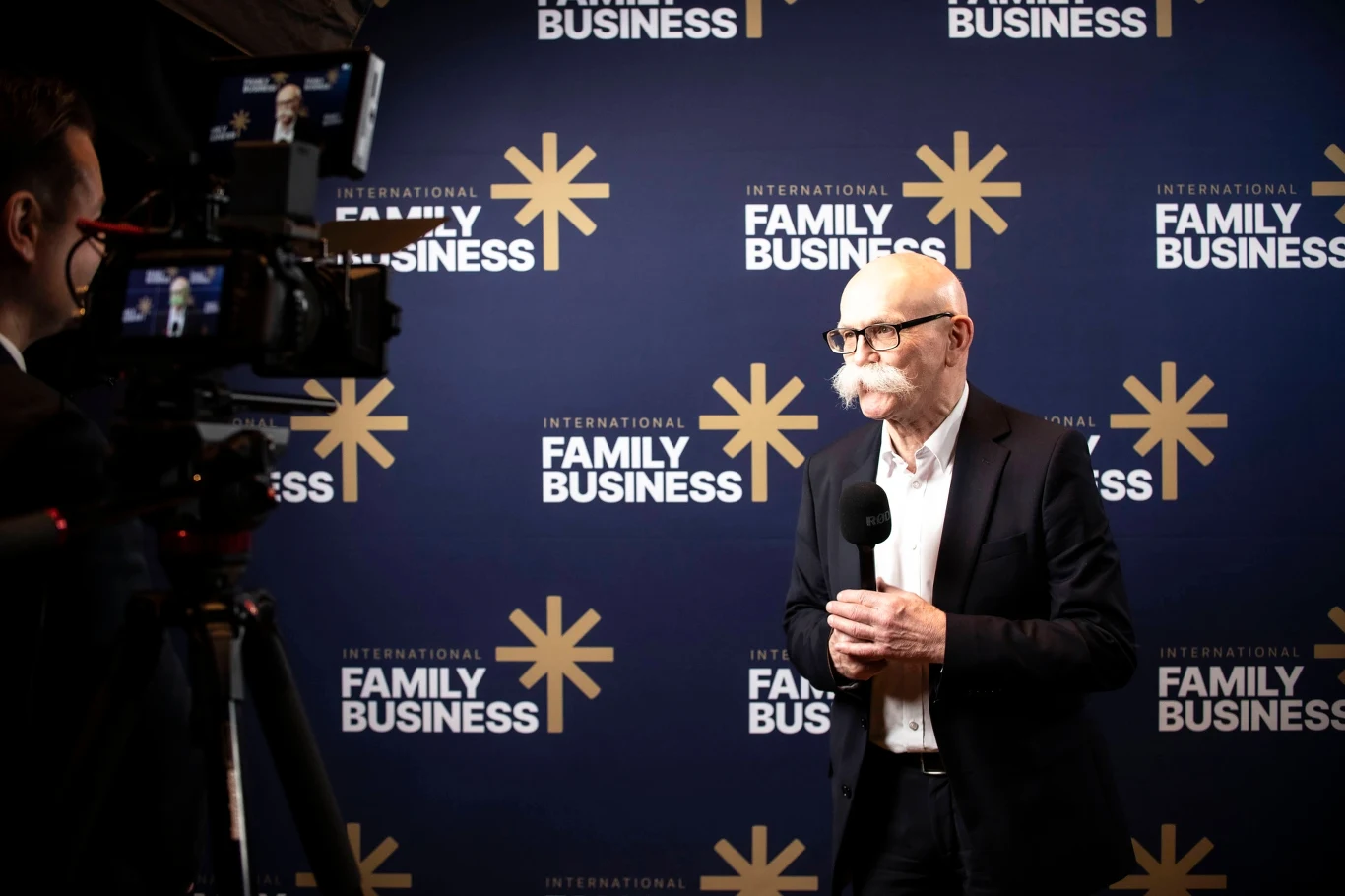 Starszy mężczyzna w garniturze, trzymający mikrofon, udziela wywiadu na tle ściany z logotypami International Family Business, obok widoczna kamera oraz operator rejestrujący rozmowę.