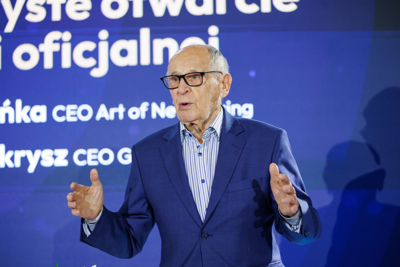 Starszy mężczyzna w okularach, ubrany w granatową marynarkę i koszulę w paski, przemawia na tle niebieskiego ekranu z napisem zawierającym nazwy stanowisk i imion organizatorów.