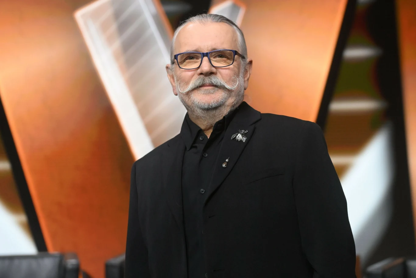 Artur Słowik został finalistą "The Voice Senior" Starszy mężczyzna z siwym wąsem i okularami ubrany w czarną marynarkę i koszulę stoi na tle rozpoznawalnego, geometrycznego tła z dużą literą W. Wygląda poważnie i pewnie.