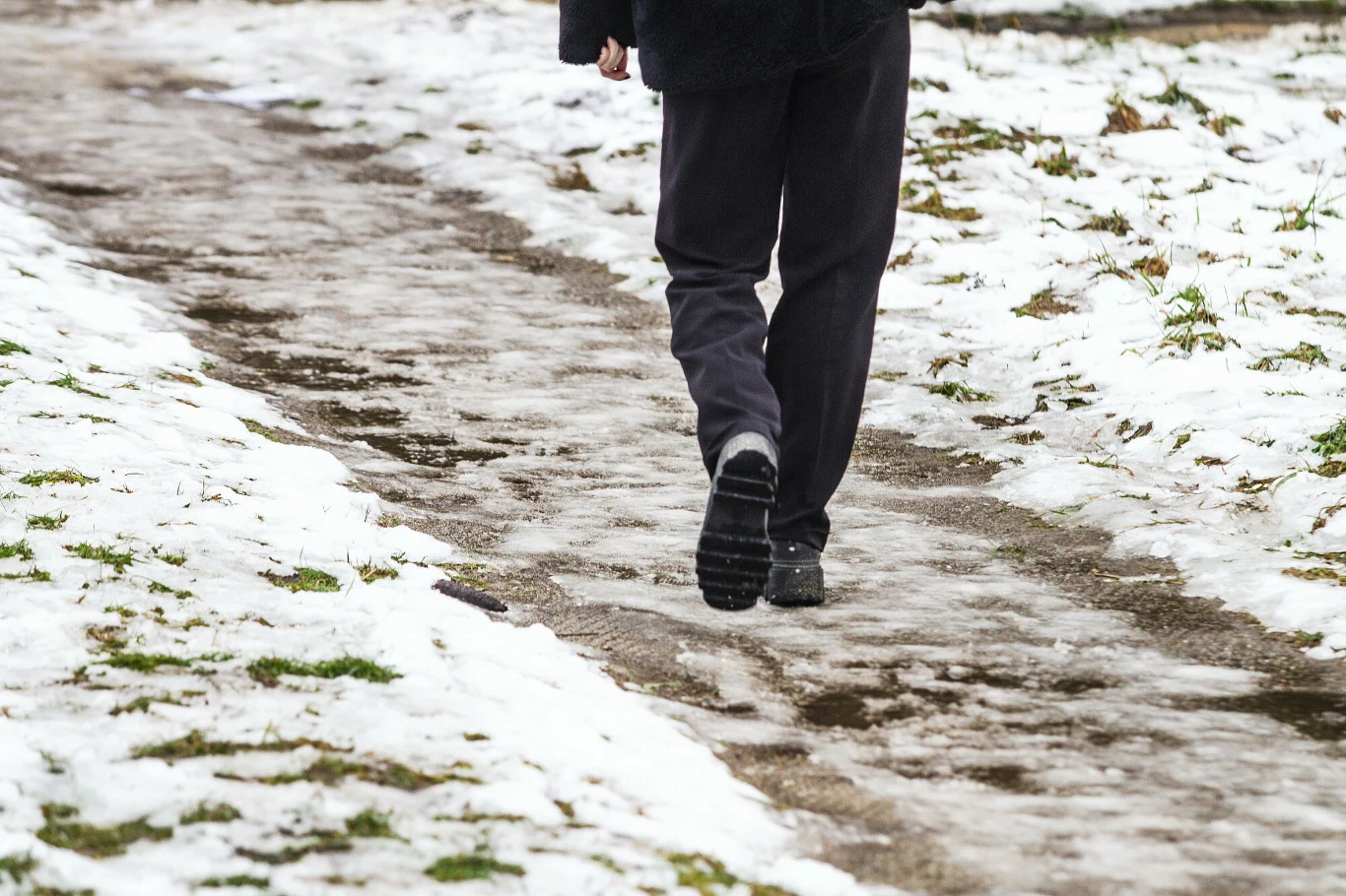 Osoba idąca po śliskiej, oblodzonej ścieżce, otoczonej topniejącym śniegiem i fragmentami trawy.