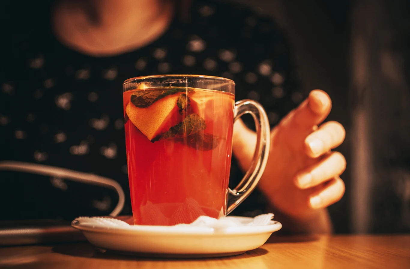 W których herbatach owocowych znajduje się najwięcej pestycydów? Szklanka gorącej herbaty z plasterkiem cytryny i ziołami na spodku, dłoń sięgająca po napój.