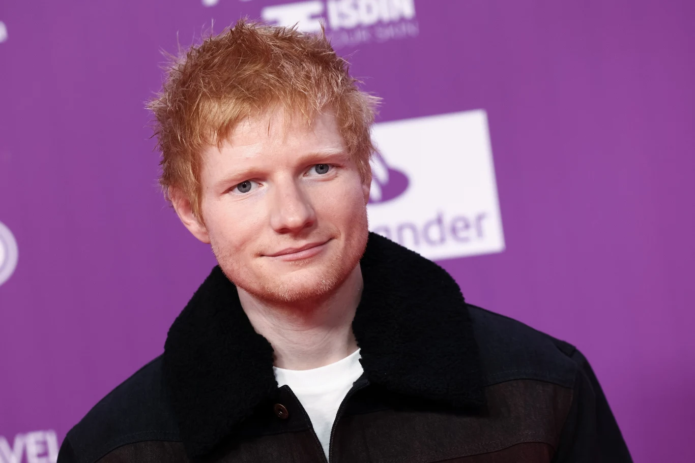 Ed Sheeran kończy 35 lat Mężczyzna z rudymi włosami i jasną cerą ubrany w ciemną kurtkę, stojący na tle fioletowej ścianki z logotypami sponsorów.