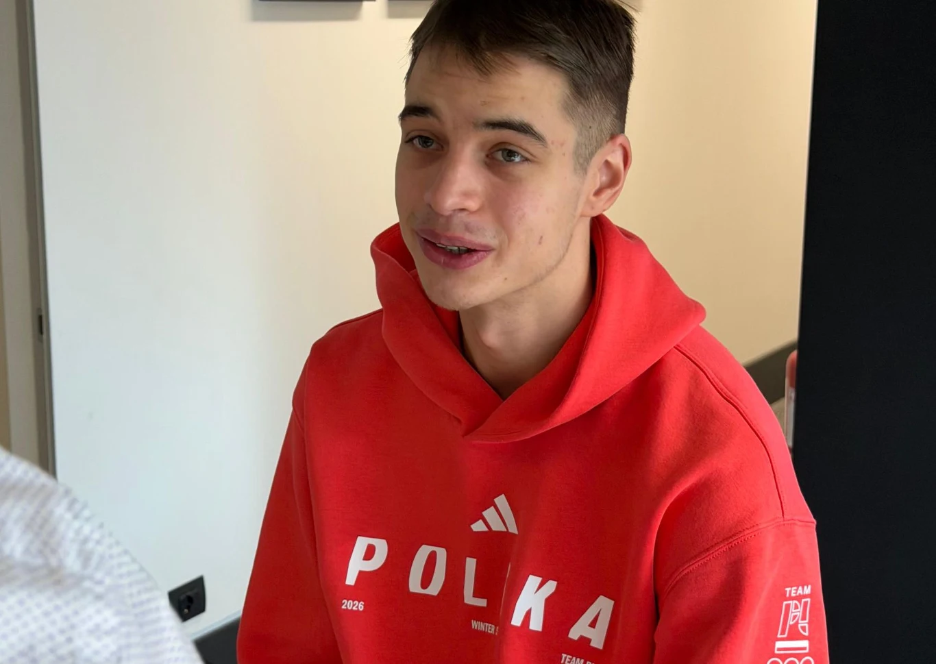 Młody mężczyzna w czerwonej bluzy z napisem 'POLKA' siedzi w jasno oświetlonym pomieszczeniu, lekko uśmiechnięty, zwrócony w stronę rozmówcy.