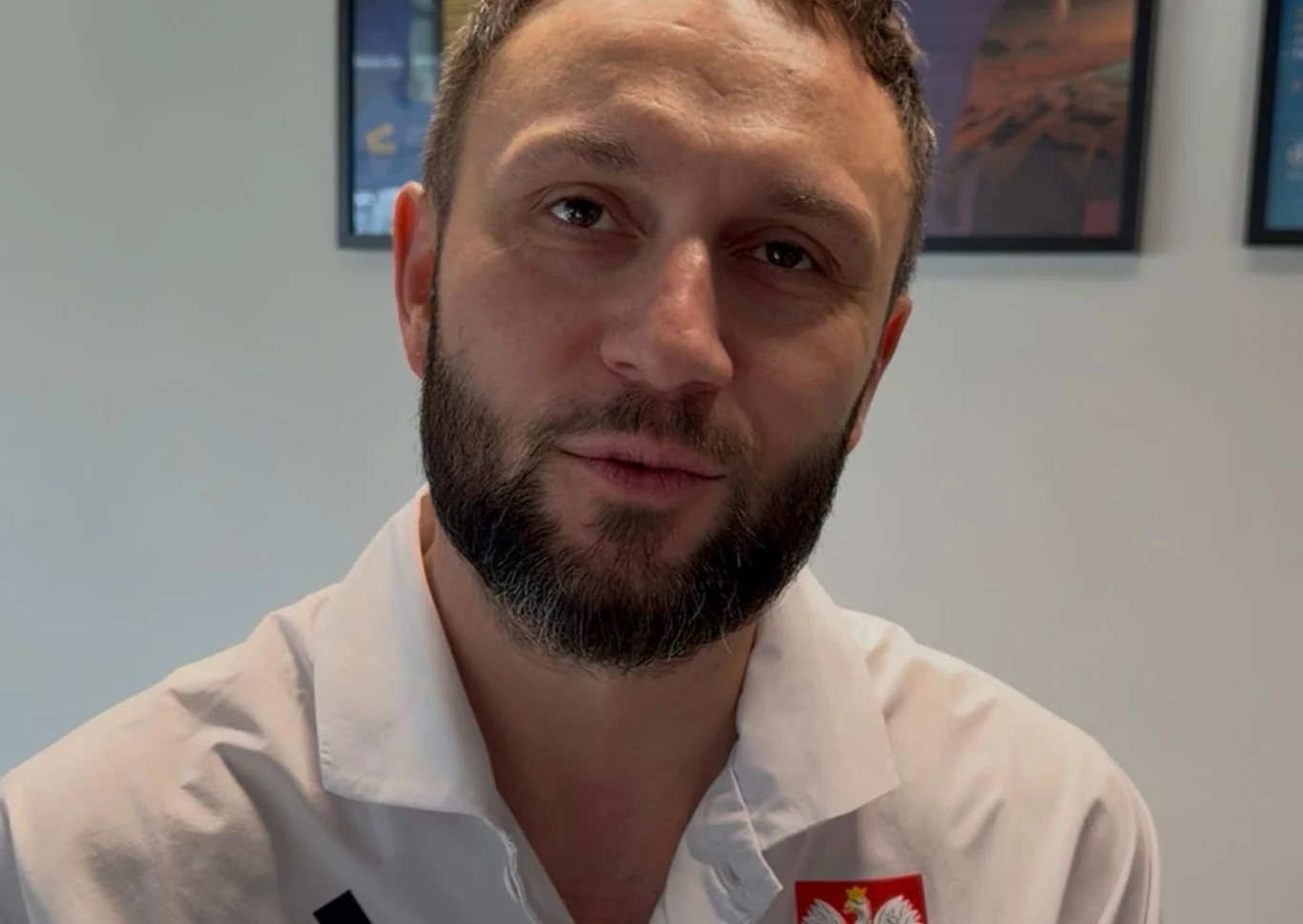 Mężczyzna z brodą w białej koszuli z godłem Polski na piersi, patrzący prosto w obiektyw. W tle fragmenty obrazów lub plakatów na ścianie.