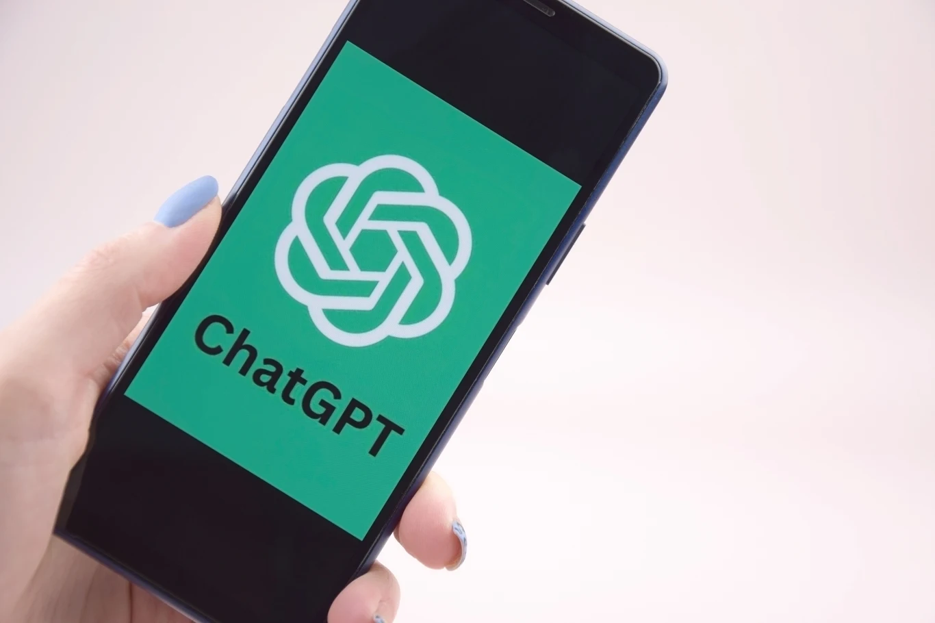 Ekran smartfona z widocznym logo i napisem ChatGPT, trzymany w ręce z niebieskim lakierem do paznokci, na jasnym tle.