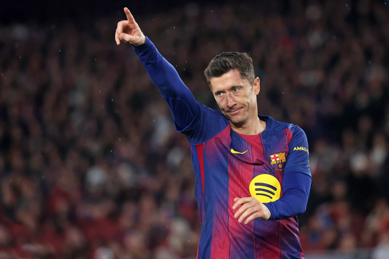 Piłkarz w stroju FC Barcelony podczas meczu na stadionie, unosi rękę i wskazuje palcem w górę, na jego twarzy widoczny jest uśmiech, w tle rozmyte sylwetki kibiców.