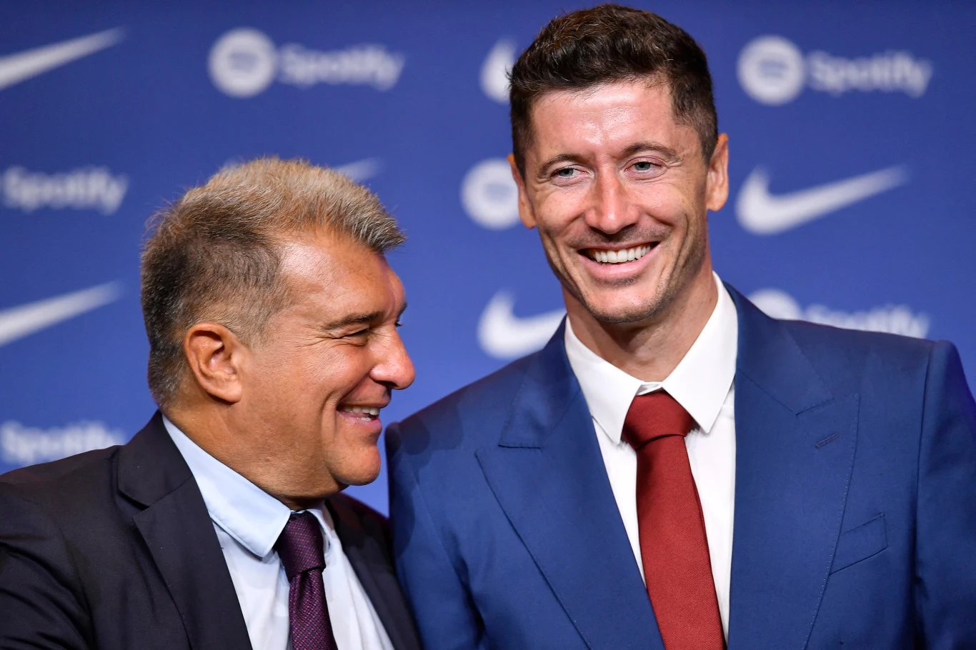 Dwaj mężczyźni w eleganckich garniturach stoją obok siebie na tle sponsorujących logotypów, uśmiechnięci, wyraźnie zadowoleni.