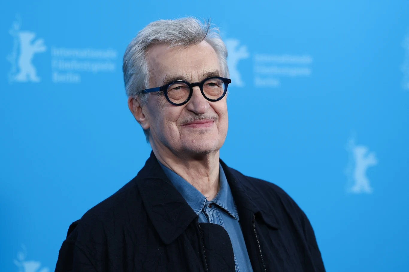 Mężczyzna w okularach o siwych włosach, ubrany w elegancką koszulę i ciemną marynarkę, stoi na tle niebieskiej ścianki z logo festiwalu filmowego.