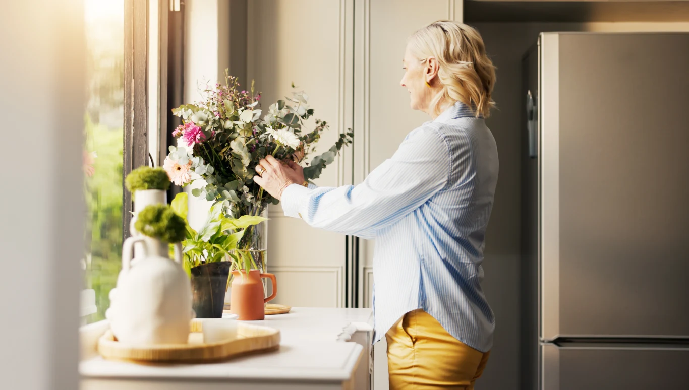 Kobieta w średnim wieku układa bukiet świeżych kwiatów stojących na kuchennym blacie w jasnej kuchni, do której wpada naturalne światło przez okno.