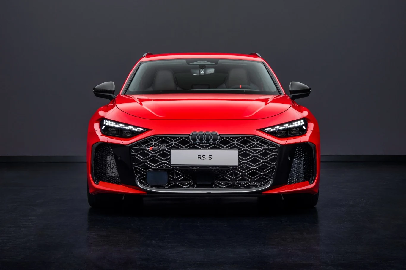 Czerwone sportowe Audi RS 5 na tle ciemnej, jednolitej ściany, widoczne bezpośrednio od przodu, z charakterystyczną dużą kratą chłodnicy, agresywnym wzornictwem reflektorów i dynamiczną sylwetką