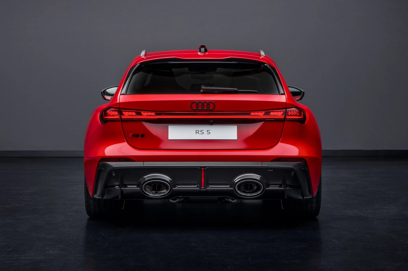 Czerwony sportowy samochód Audi RS 5 widoczny od tyłu, wyraźnie podświetlone tylne światła, masywny zderzak z dwoma dużymi końcówkami wydechu i emblematem modelu, tle ciemne neutralne.