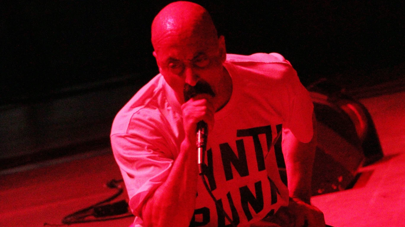 Łysy mężczyzna w jasnej koszulce z napisem, śpiewający lub krzyczący do mikrofonu podczas koncertu, oświetlenie w intensywnym czerwonym kolorze, widoczne wyraziste emocje.