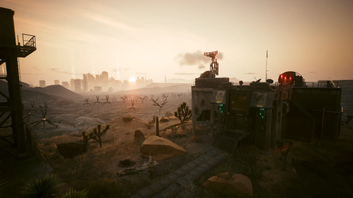 Opustoszały krajobraz pustynny z industrialnym budynkiem po prawej stronie, otoczony suchymi roślinami i kamieniami, w tle ogrodzenie z drutu kolczastego oraz miasto na horyzoncie, oświetlone zachodzącym słońcem.