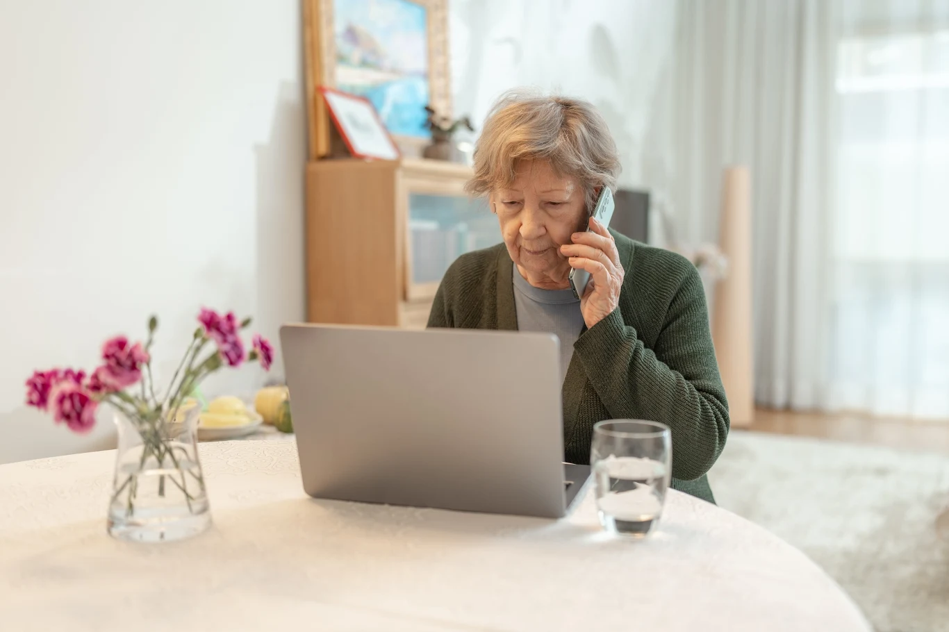 Telefonicznych oszustwom ulegają dziś nie tylko seniorzy, jednak to oni powinni być najbardziej czujni Starsza kobieta skupiona na rozmowie przez telefon, siedzi przy stole z laptopem i szklanką wody. Na stole stoi również wazon z różowymi kwiatami, w tle widoczna komoda i jasne zasłony.