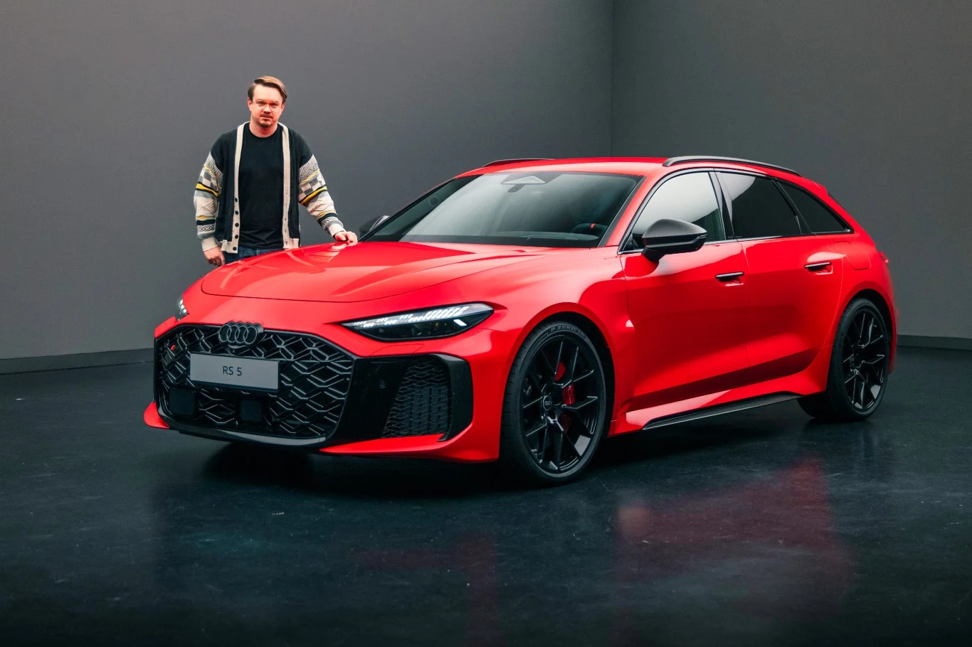 Czerwony sportowy samochód Audi RS 6 ustawiony w studiu na tle szarej ściany. Obok pojazdu stoi mężczyzna ubrany w casualową kurtkę i ciemne spodnie.