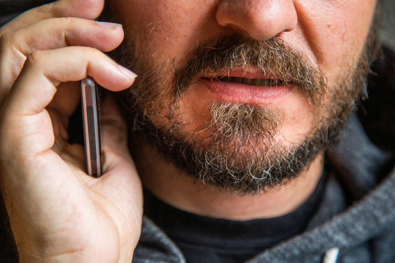 Oszuści wykorzystują połączenia telefoniczne w celu wyłudzenia danych oraz pieniędzy Zbliżenie na dolną część twarzy mężczyzny z brodą w szarej bluzie, trzymającego telefon przy uchu, jakby prowadził rozmowę.