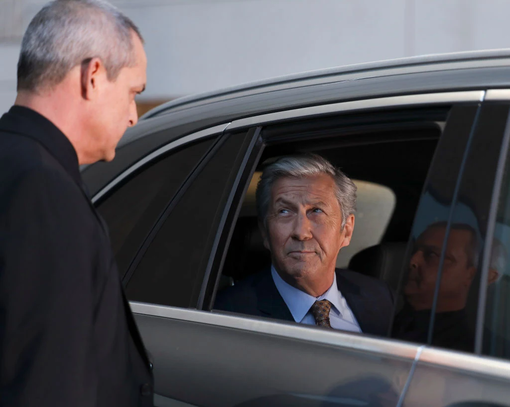 Mężczyzna siedzi na tylnym siedzeniu samochodu, spogląda przez otwarte okno na stojącego na zewnątrz innego mężczyznę w ciemnym garniturze.