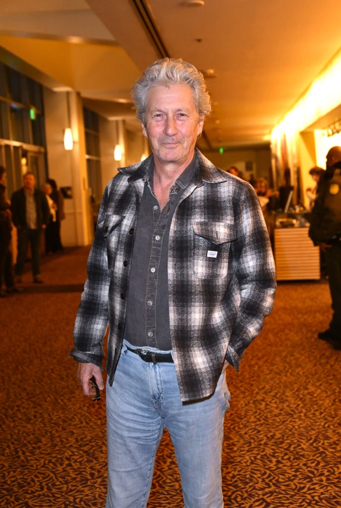 Starszy mężczyzna z siwymi włosami ubrany w kraciastą koszulę i dżinsy stoi w przestronnym, dobrze oświetlonym korytarzu. W tle widoczne są osoby oraz elementy wnętrza, takie jak dywan i oświetlenie sufitowe.