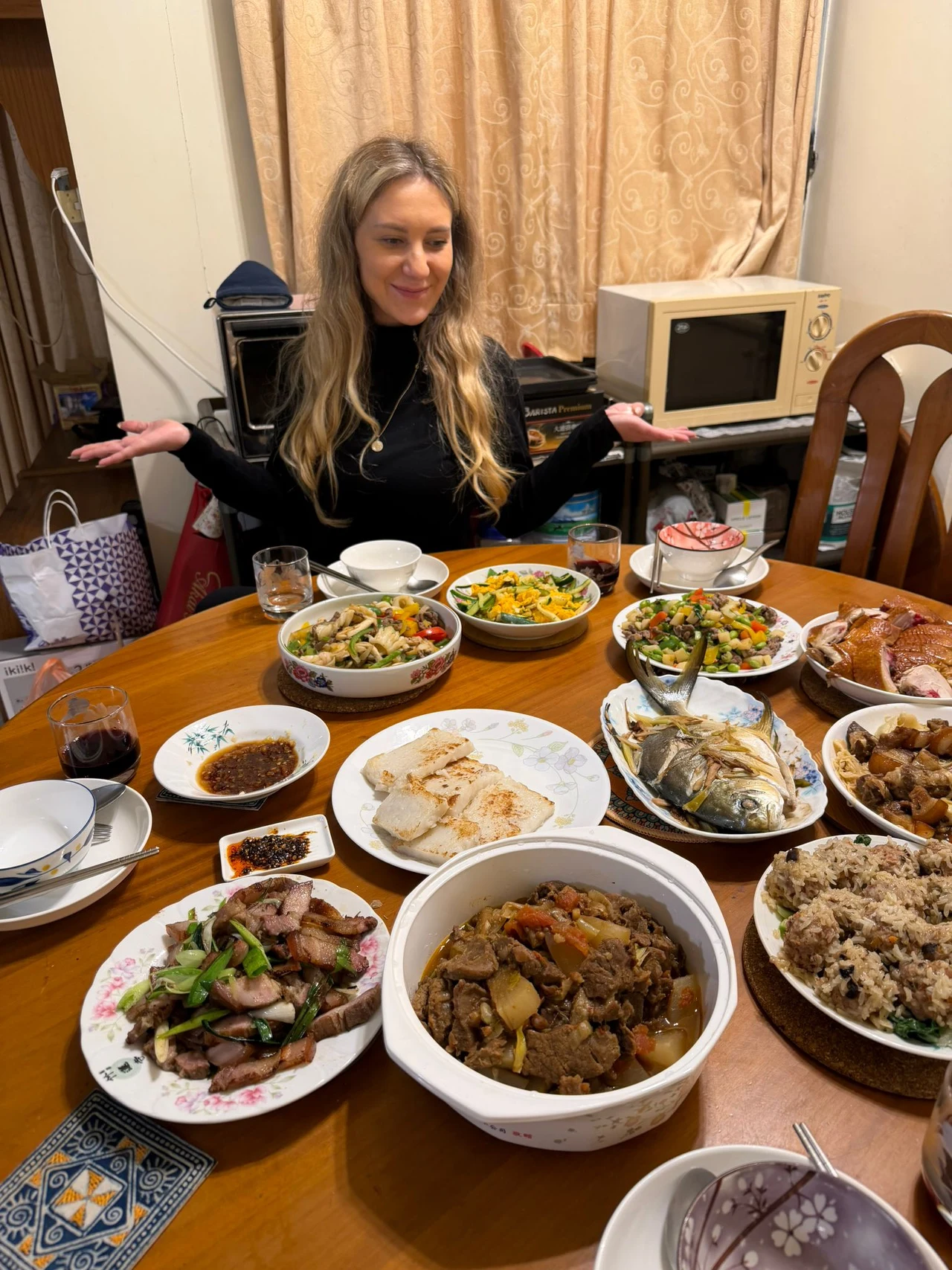 Kolacja (prawie jak) Wigilia. Tylko jeszcze bardziej “na bogato” Kobieta z jasnymi włosami siedzi przy stole zastawionym różnorodnymi potrawami kuchni azjatyckiej i europejskiej, wśród dań widoczne są ryby, mięsa, sałatki oraz ryż. W tle znajduje się mikrofalówka, zasłonięte okno oraz krzesła.