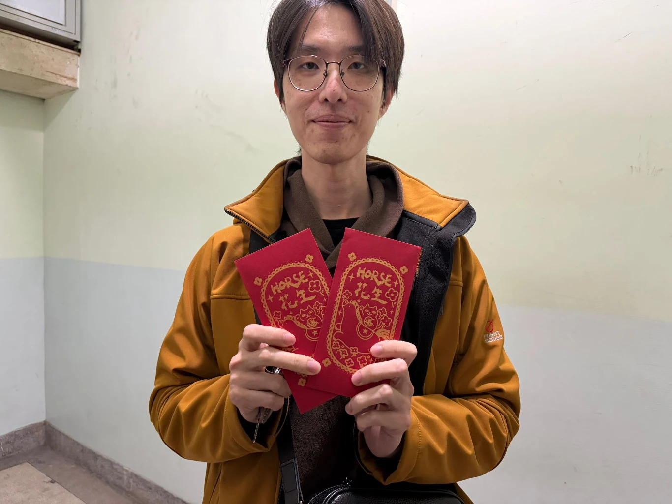 Czerwone koperty Mężczyzna w okularach i pomarańczowej kurtce trzyma w dłoniach dwie czerwone koperty z ozdobnym złotym nadrukiem oraz napisem HORSE, stojąc na tle jasnej, pustej ściany.