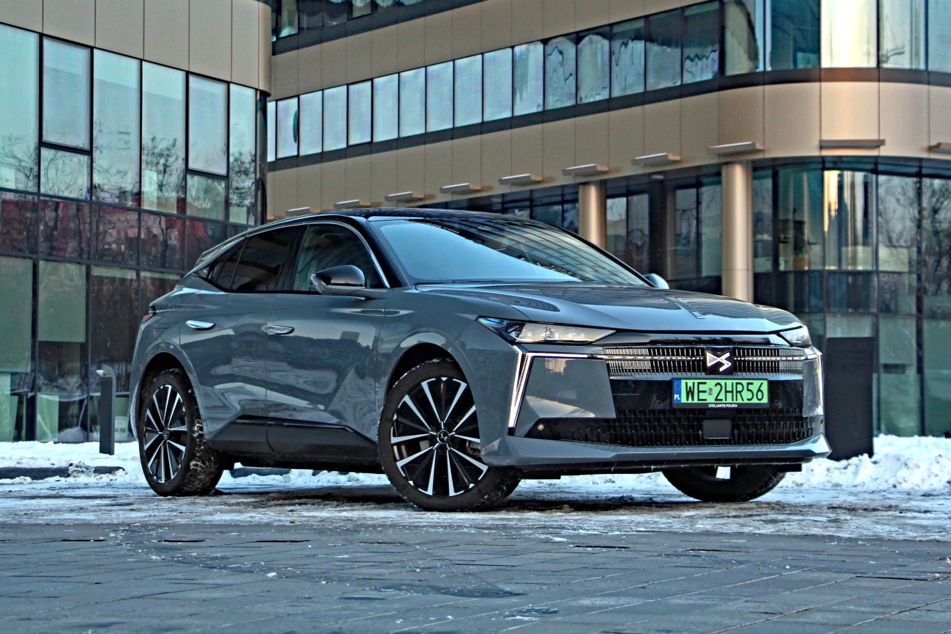 DS No4 E-Tense, czyli zmodernizowany francuski kompakt w wersji elektrycznej Szary, nowoczesny samochód osobowy z czarnymi felgami stoi zaparkowany na tle przeszklonego budynku biurowego, na chodniku z resztkami śniegu.