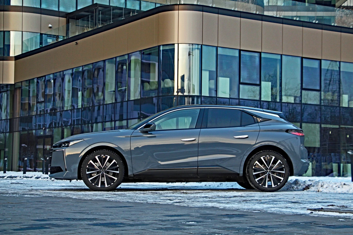 DS No4 nadal zwraca uwagę swoją sylwetką Szary, nowoczesny samochód osobowy zaparkowany na tle przeszklonego budynku biurowego z elementami złotego wykończenia, na nawierzchni pokrytej śniegiem.