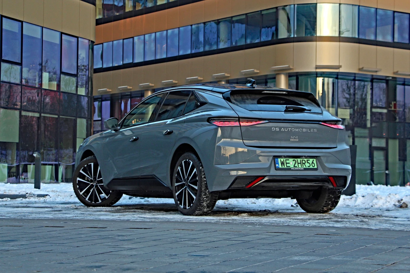 Duży napis DS AUTOMOBILES pozwala łatwo odróżnić No4 po liftingu Srebrny samochód marki DS Automobiles zaparkowany na tle nowoczesnego, szklanego budynku. Na nawierzchni widoczny śnieg, auto stoi na chodniku, a jego nowoczesny design i dynamiczne linie przyciągają uwagę.