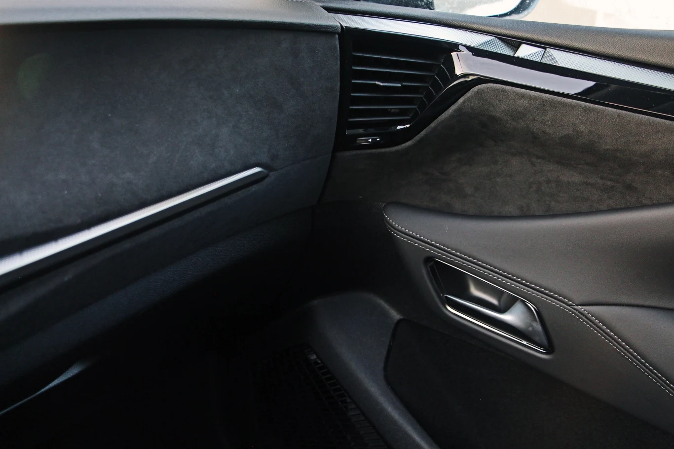 Deska rozdzielcza płynnie przechodzi w panel drzwi, a wrażenie jednolitości obu tych elementów wzmagają nawiewy, które znajdują się na drzwiach. Połączenie alcantary i wysokiej jakości skóry nadają wnętrzu DS No4 trochę szlachetnego klimatu Wnętrze samochodu z widocznym fragmentem drzwi, otworem na klamkę oraz czarną deską rozdzielczą i eleganckim wykończeniem z przeszyciami.