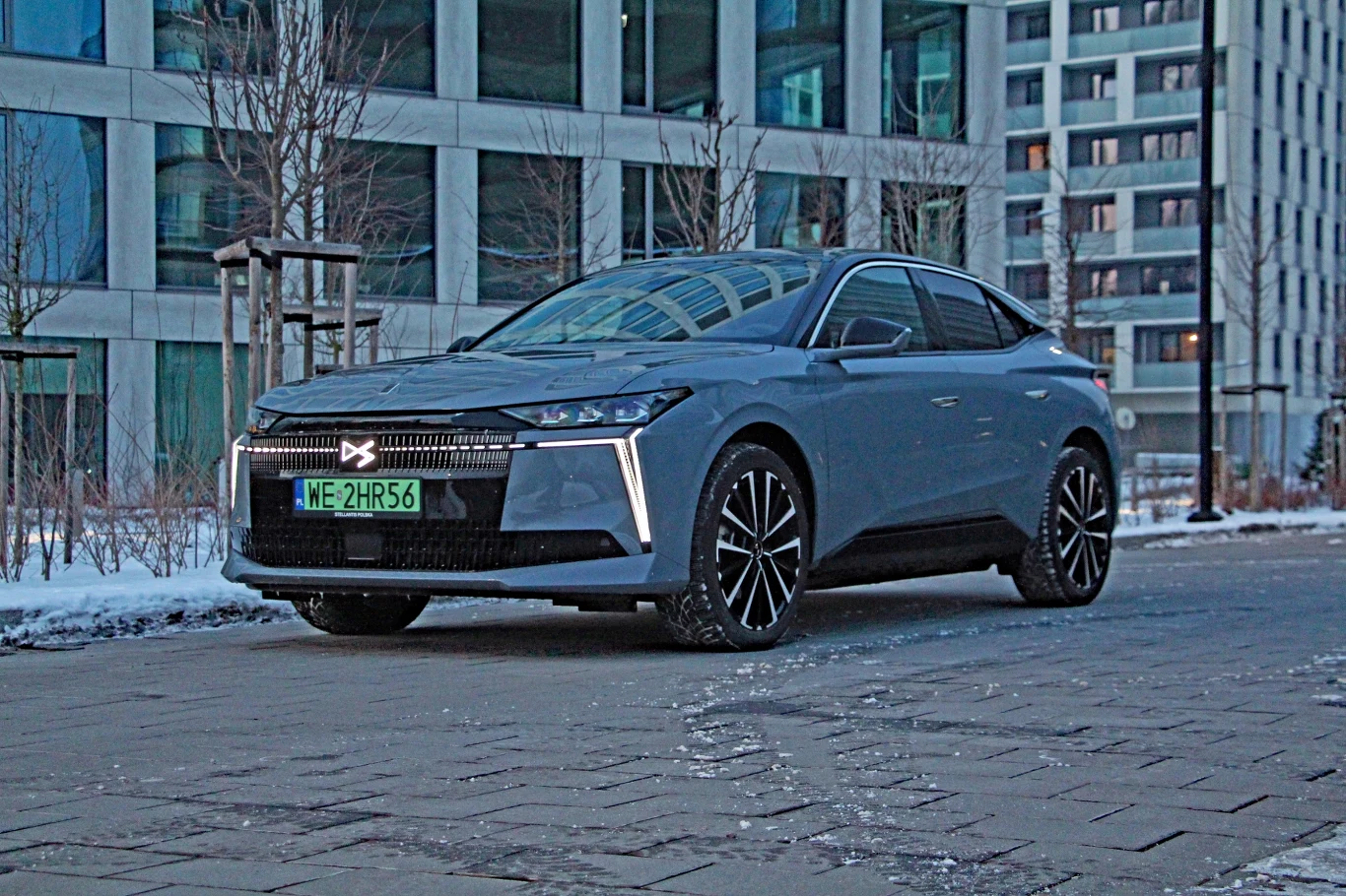 DS No4 E-Tense Nowoczesny, srebrny samochód elektryczny zaparkowany na tle szklanych biurowców w zimowej scenerii. Auto wyróżnia się stylowymi felgami i charakterystycznym oświetleniem LED.