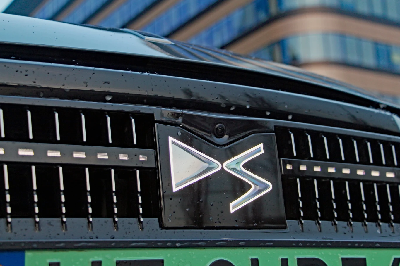 DS No4 E-Tense Zbliżenie na przedni grill samochodu marki DS Automobiles z widocznym logotypem oraz kroplami deszczu na karoserii, w tle fragment nowoczesnego biurowca.