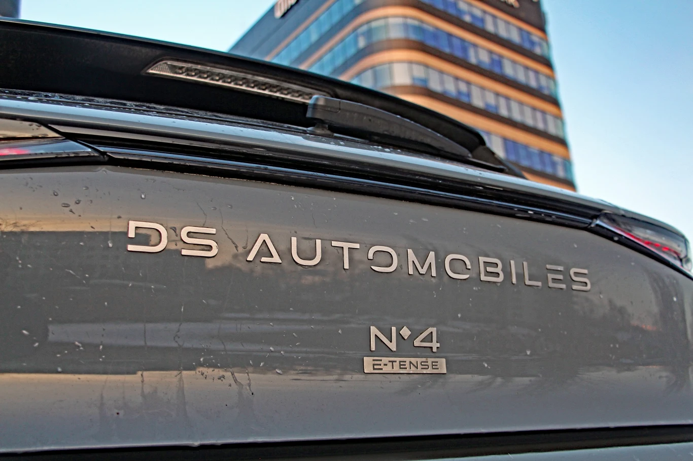 DS No4 E-Tense Tył samochodu marki DS Automobiles z wyraźnie widocznym emblematem modelu N°4 E-Tense, w tle nowoczesny biurowiec.