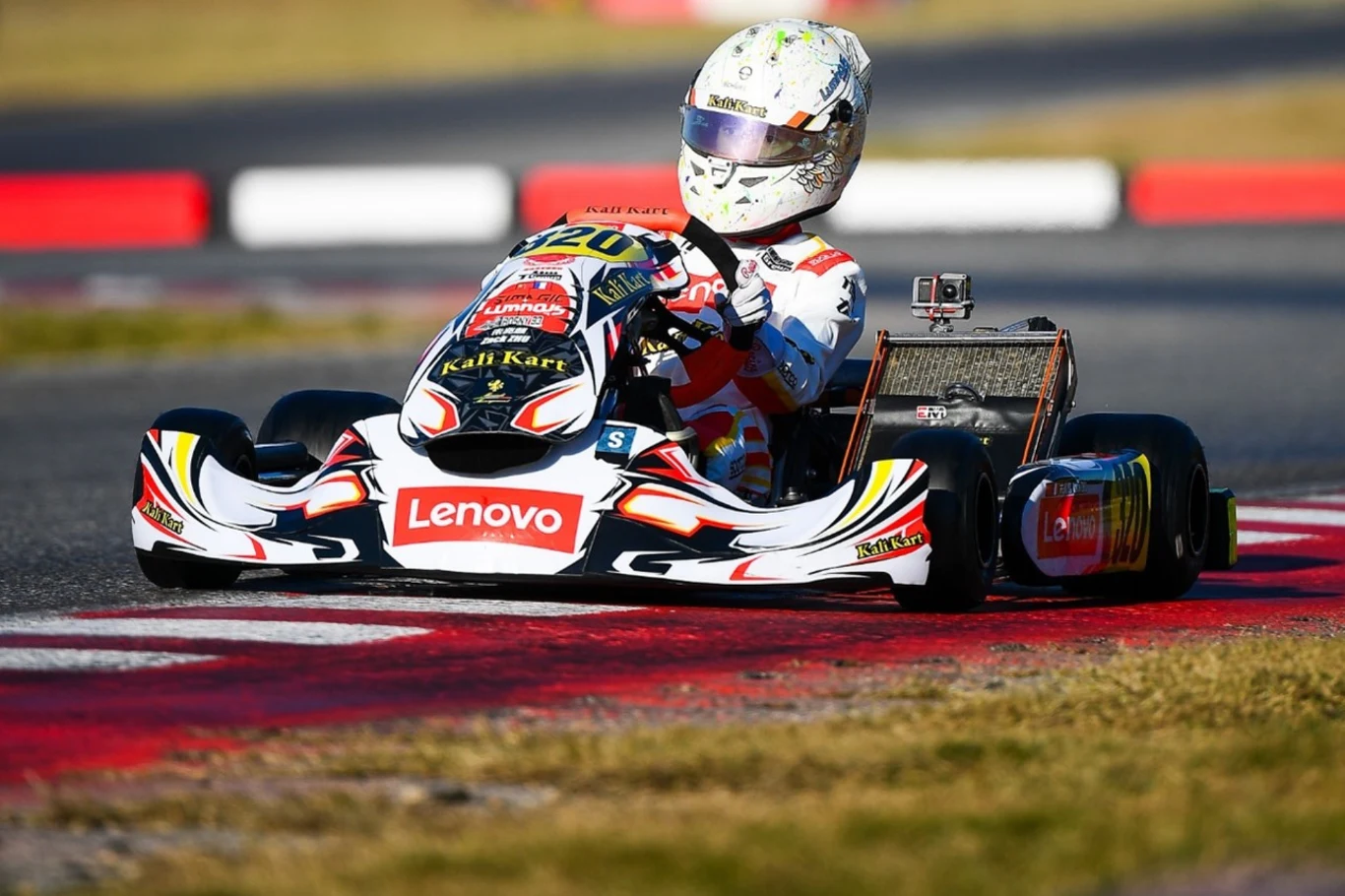 Kierowca w białym kasku prowadzi kolorowy gokart po torze wyścigowym, na pojeździe widoczne są logotypy sponsorów, w tle asfaltowa nawierzchnia toru i fragment pobocza z trawą.