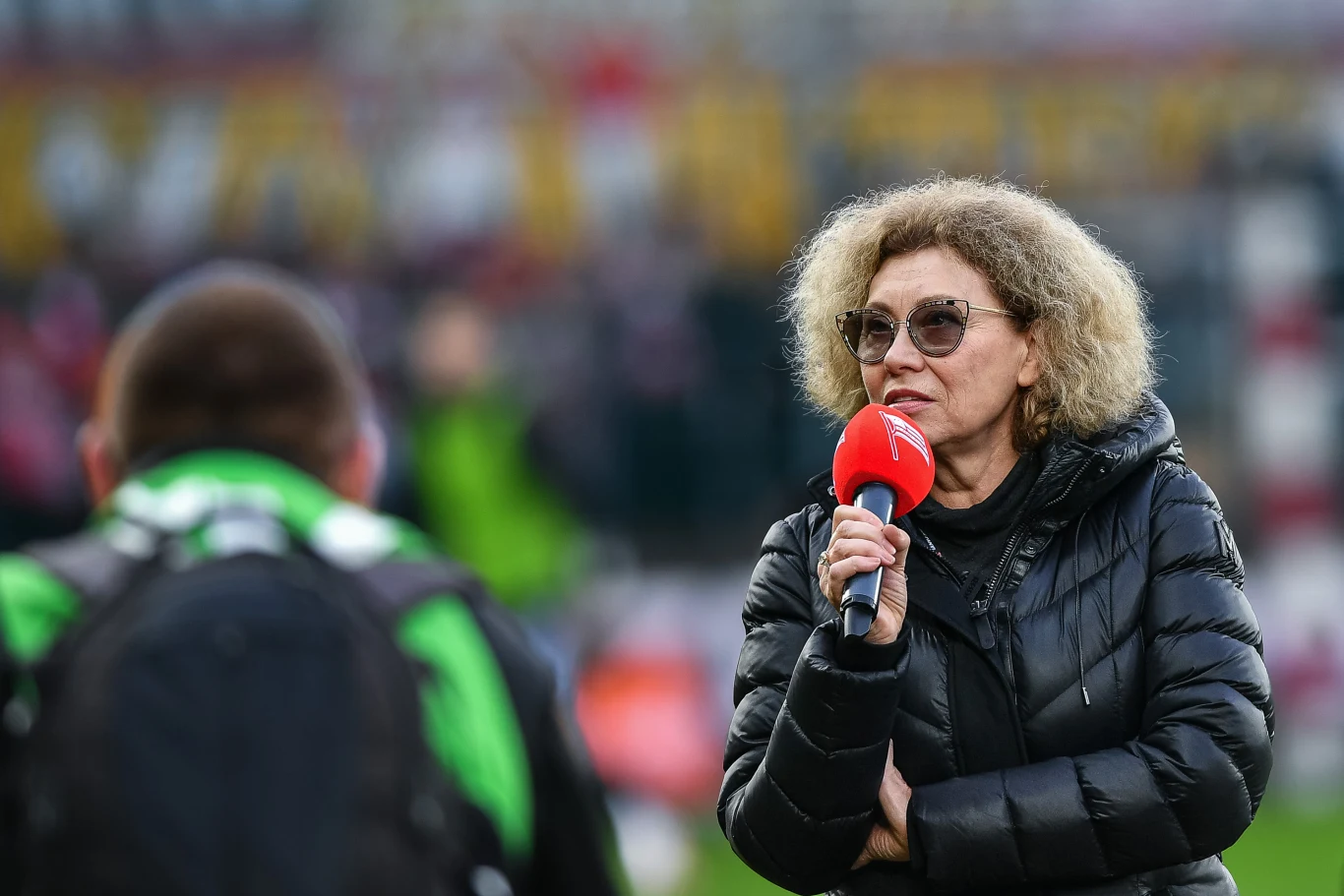 Elżbieta Filipiak Kobieta w okularach przeciwsłonecznych trzymająca mikrofon z czerwonym oznaczeniem udziela wywiadu na tle rozmytego stadionu, w kadrze widoczna sylwetka innej osoby skierowanej plecami do aparatu.