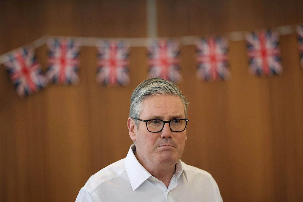 Mężczyzna w okularach i białej koszuli stoi na tle brązowej ściany, nad którą widoczne są girlandy z małych brytyjskich flag.