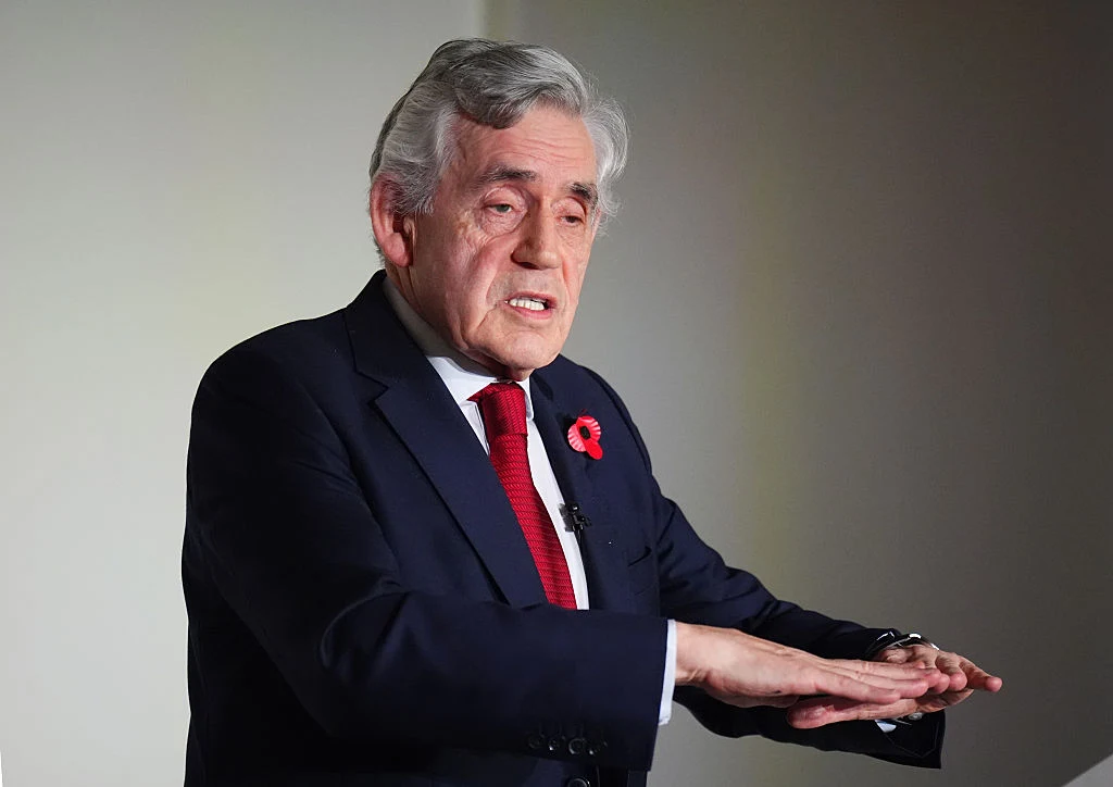 Starszy mężczyzna w garniturze i czerwonym krawacie, z przypiętym kwiatkiem do klapy, wykonuje gest rękami, prawdopodobnie wyjaśniając coś lub przemawiając.