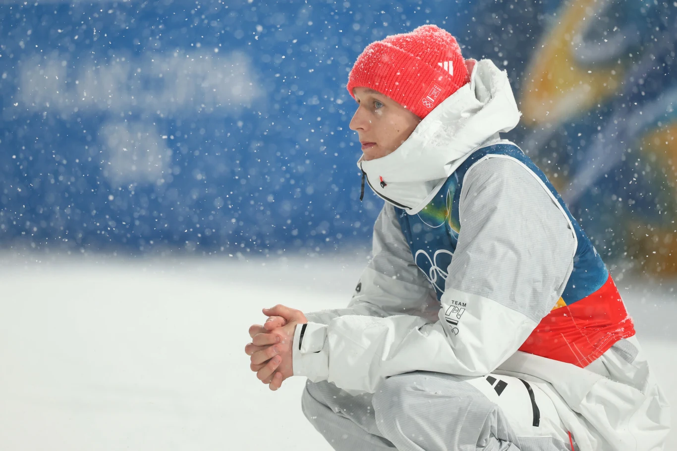 Paweł Wąsek Osoba w stroju sportowym i czerwonej czapce siedząca na śniegu podczas opadów śniegu, w tle rozmazane logo oraz niebieski baner z motywem sportowym.