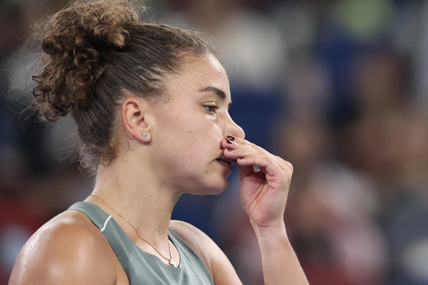 Jasmine Paolini Młoda kobieta o kręconych włosach spiętych w kucyk stoi bokiem, trzymając dłoń przy ustach, zamyślona. Ubrana w sportowy strój, w tle nieostre sylwetki oraz rozmyte kolory sugerujące obecność publiczności.